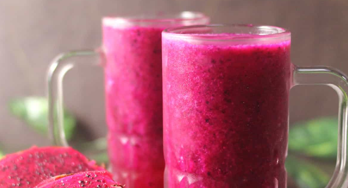 A glass of vibrant pink dragon fruit juice garnished with fresh dragon fruit slices, mint leaves, and lime, served alongside fresh tropical fruits.