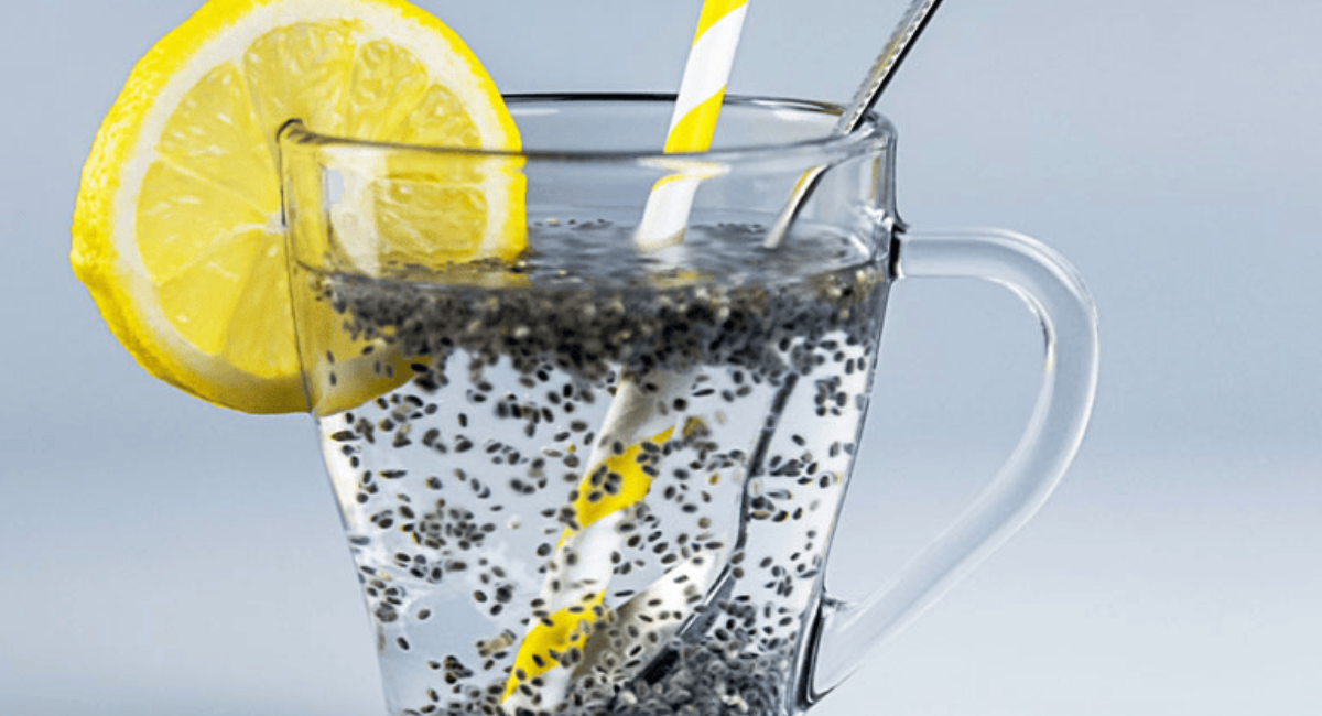 A glass bottle filled with chia seed water, lemon slices, and soaked chia seeds, alongside fresh lemon wedges and chia seeds on a wooden surface.